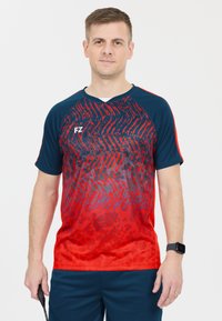 Röd och marinblå sportskjorta med raglanärm, som har ett texturerat vågmönster. Tillverkad av lättviktig tyg, kombinerad med marinblå shorts.