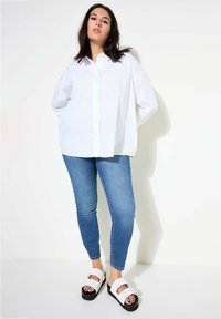 Camicia bianca oversize con dettagli decorativi sulle spalle, abbinata a jeans skinny azzurri e sandali neri con piattaforma e strisce bianche.