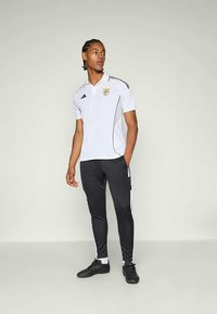 Hvit polo-skjorte med svarte detaljer og emblem, sammen med svarte treningsbukser med hvite sidestriper og svarte sportsko.