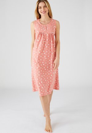 Donna sorridente, che cammina a piedi nudi, indossa un abito rosa senza maniche fino al ginocchio con motivo floreale bianco su sfondo uniforme.