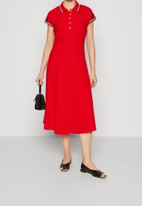 Rode polojurk met korte mouwen, witte en marineblauwe gestreepte kraag, en knopen aan de voorkant; gecombineerd met zwarte ballerina's met gouden accenten.