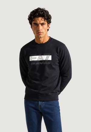 Mand med krøllet hår iført sort EA7 Emporio Armani sweatshirt og blå jeans, stående mod en ensfarvet lys baggrund.
