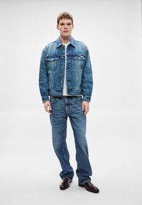 Μπουφάν και τζιν παντελόνι σε ανοιχτό μπλε χρώμα, με κουμπιά στην μπροστινή πλευρά, τσέπες στο στήθος και ίσια γραμμή στο πόδι. Συνοδεύεται από σκούρα καφέ παπούτσια.