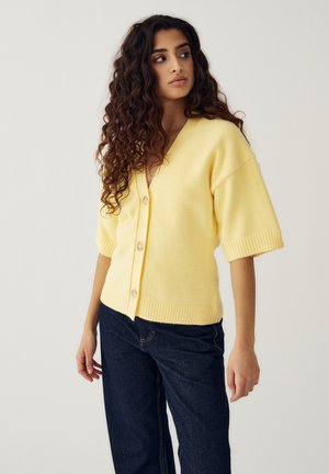 Donna con capelli lunghi e ricci che indossa un cardigan giallo con bottoni e maniche corte, e jeans blu scuro, in piedi davanti a uno sfondo uniforme.