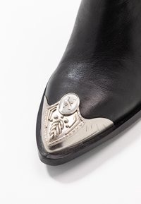 Černá kožená cowboyská bota s špičatou špičkou, zdobená stříbrnými kovovými prvky a podrobnými rytinami na špičce.