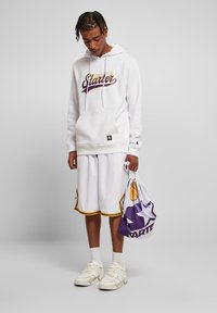 Haut à capuche blanc avec le logo "Starter" en violet et or, associé à un short blanc avec une bordure jaune. Tient un sac en mesh en violet et blanc.