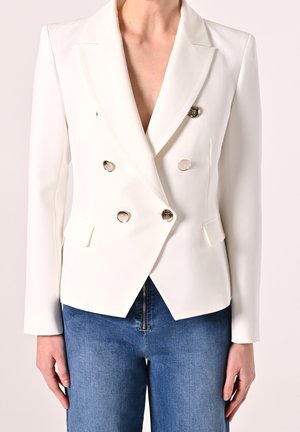 Blazer blanc croisé à boutons argentés porté sur un jean bleu, avec un détail de fermeture éclair à l'avant du jean.