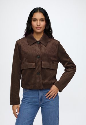 Femme aux longs cheveux foncés portant une veste boutonnée marron foncé avec de grandes poches et un jean bleu sur un fond clair uni.