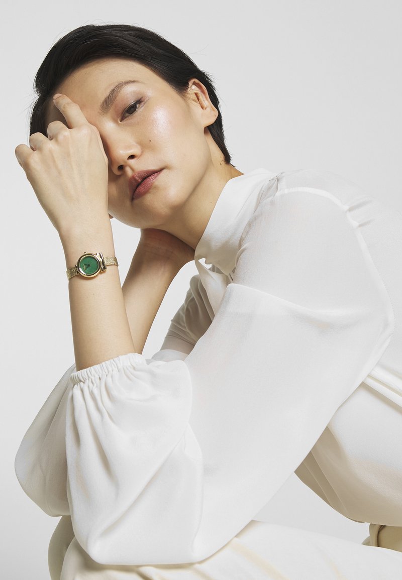 Montre-bracelet en or avec un cadran vert, un bracelet texturé et un visage circulaire. Le modèle porte une blouse blanche à manches volumineuses sur un fond neutre.