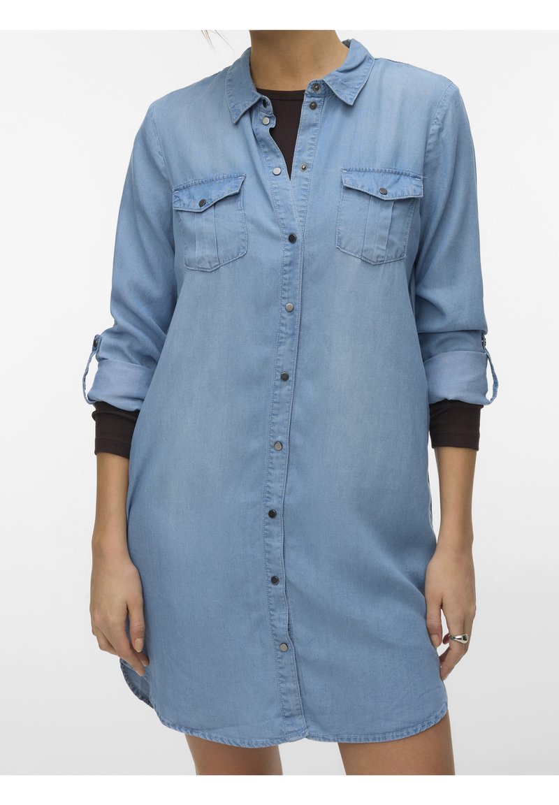 Femme portant une robe-chemise en jean bleu clair boutonnée avec des manches retroussées et un t-shirt à manches longues foncé en dessous.