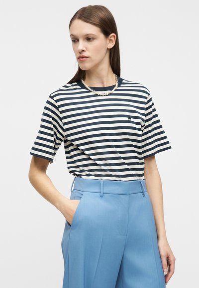 Γυναίκα με ίσια καστανά μαλλιά φορά μπλε σκούρη και λευκή ριγέ κοντομάνικη μπλούζα, μαργαριταρένιο κολιέ και ψηλόμεσα ανοιχτό μπλε παντελόνια, με το ένα χέρι στην τσέπη.