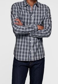 Szara i czarna koszula w kratę zapinana na guziki, wykonana z materiału o miękkiej teksturze. Posiada długie rękawy i kołnierzyk, noszona z ciemnymi dżinsami.