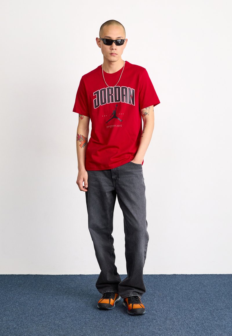 Rotes Baumwoll-T-Shirt mit "JORDAN"-Grafik, kombiniert mit dunkelgrauen Jeans und schwarz-orangefarbenen Sneakers. Das Modell trägt Sonnenbrillen und eine Halskette.
