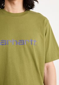 Olivgrünes Baumwoll-T-Shirt mit kurzen Ärmeln, mit einem großen blauen grafischen Text "carhartt" über der Brust.