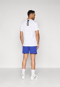 Λευκό καμουφλάζ κοντομάνικο T-shirt με μαύρη ετικέτα "BORG" στην πλάτη, συνδυασμένο με μπλε βερμούδες και λευκά αθλητικά παπούτσια.