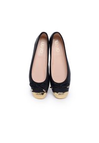 Zapatos planos negros con una textura suave, con punta redondeada, puntera dorada y un pequeño detalle de lazo en el empeine. Forro interior suave.