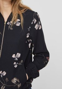 Svart bomberjacka med blommönster i rosa och gyllene detaljer, med zip fram, elastiska ärmslut och avslappnad passform.