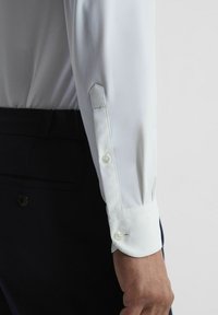 Vit långärmad skjorta med knappmanchetter, slät textur, slim fit-design och minimalism, mot en kontrasterande mörk bakgrund.