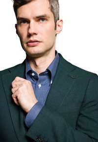 Zielona marynarka o fakturze noszona na niebieskiej koszuli z guzikami, z charakterystycznym kołnierzykiem i białymi guzikami. Marynarka ma mały czarny akcent.