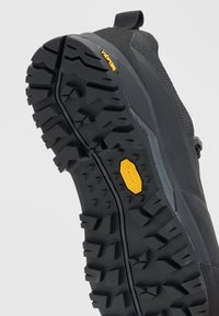 Черна подметка на туристическа обувка с дълбоки грайфери и жълто лого Vibram на сив фон.