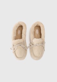 Chaussons beiges avec une doublure douce et pelucheuse, un bout rond et une semelle plate. Ils présentent un détail en dentelle et un design simple, sans embellissement.