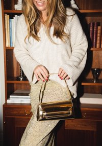 Złota metaliczna torebka o smukłym kształcie i krótkim uchwycie, zestawiona z błyszczącą złotą spódnicą i białym swetrem w prążki.