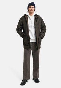 Chaqueta exterior marrón oscuro con dos bolsillos frontales, combinada con una sudadera blanca y pantalones anchos grises. Zapatos negros y un gorro azul marino completan el look.