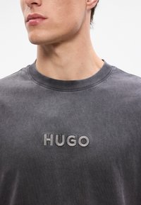Graues Baumwoll-T-Shirt mit Rundhalsausschnitt, das auf der Brust ein geprägtes "HUGO"-Logo in einer helleren Grauschattierung zeigt. Weicher Stoff.