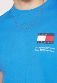 Modré bavlněné tričko s černým, bílým a červeným obdélníkovým logem s textem "TOMMY JEANS". Také je viditelný stříbrný řetízek.