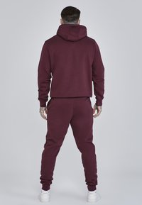 Tuta maroon con cappuccio, caratterizzata da polsini e vita a costine, mostrata da dietro. Realizzata in tessuto morbido, design aderente. Sneakers bianche.