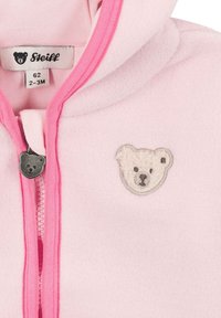 Rosa Fleecejacke mit Kapuze. Sie verfügt über einen Reißverschluss, pinkfarbene Akzente und eine graue Bärenkopf-Stickerei auf der Brust. Markenetikett innen sichtbar.