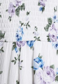 Vit tyg med små blå och lila blommönster, med en samlad shirring-detalj högst upp och mjuka veck nedanför.