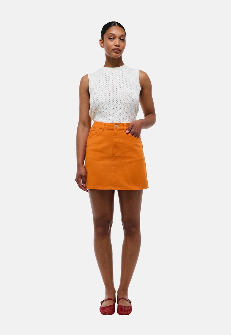 Haut sans manches en tricot blanc associé à une jupe courte évasée orange. La jupe possède un bouton à l'avant, une texture lisse et des poches latérales.