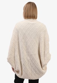 Vrouw met schouderlang haar, gekleed in een crèmekleurige gebreide poncho met een ruitpatroon, staand met handen in de zakken, weg van de camera, tegen een witte achtergrond.