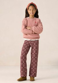 Pull en polaire rose avec un petit emblème de cœur rouge, associé à un pantalon large à motif bordeaux et des bottines beiges.