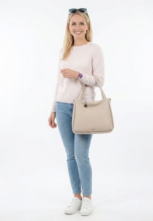 Donna che indossa un maglione rosa chiaro e jeans blu, con una borsa beige in mano, occhiali da sole sulla testa e sneakers bianche, sorridente su sfondo bianco.