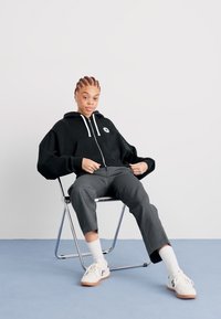 Black oversized hoodie with white drawstrings and logo, paired with grey tapered pants and white socks, sitting on a folding chair.