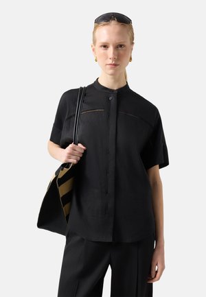 Frau trägt schwarze Hemdbluse und Hose, trägt schwarze und gelb gestreifte Tasche über der Schulter, mit Sonnenbrille auf dem Kopf.