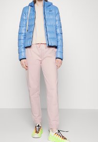 Jasnoniebieska kurtka puchowa z poziomym pikowaniem i kapturem, w połączeniu z różowymi dresami oraz intensywnie zielonymi trampkami z czarnymi akcentami.