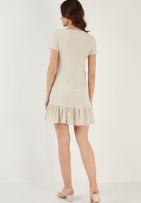 Beige jurk met korte mouwen van een gestructureerde stof, voorzien van een flonkerende volantszoom. Eenvoudig ontwerp met een ronde hals en een lengte boven de knie.