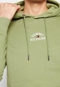 Huvtröja i ljusgrön bomull med vit "TOMMY HILFIGER"-logotyp och silverfärgade detaljer på snörningarna. Slät yta.
