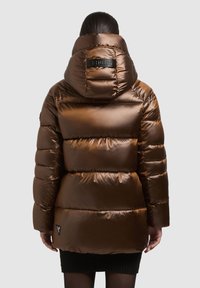 Brūna pūkaina jaka ar spīdīgu faktūru, lielu kapuci un šūtu dizainu. Aizmugurē redzama zīmola etiķete un melna apakšēja apdare.