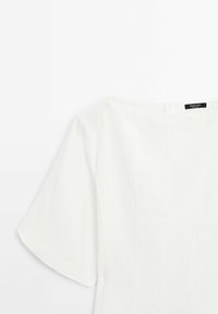 Top blanc à manches courtes avec manches larges et détails de couture, avec un col rond et une fermeture éclair au dos.