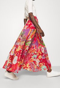 Person, die in einem maxi Rock mit roten und lila Blumenmuster, einem weißen Kurzarmtop und weißen Sneakers geht, mit einem sichtbaren Tattoo am Unterarm.