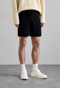Sorte shorts med elastisk linning og snor, parret med en beige strikket sweater og hvide sneakers. Tykkere hvide sokker synlige.