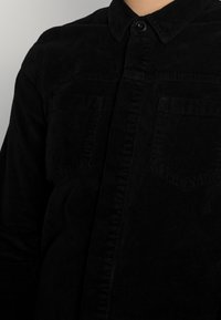 Chemise noire en velours côtelé avec un devant boutonné, un col pointu et deux poches poitrine à coupe droite avec une texture côtelée distinctive.