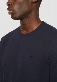 Dunkelblauer, strukturiert Pullover mit Rundhalsausschnitt, der ein tonales "BOSS"-Logo auf der Brust hat. Das Material wirkt weich und atmungsaktiv.
