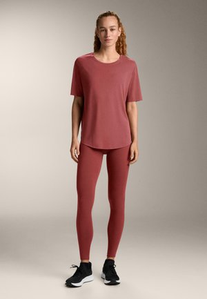 Czerwona koszulka z krótkim rękawem i zaokrąglonym dołem, zestawiona z dopasowanymi legginsami. Miękka tkanina ma gładką fakturę. Czarnie buty sportowe dopełniają stylizację.