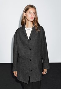 Czarny, dopasowany płaszcz w odcieniu ciemnoszarym z wyciętym kołnierzem, pięcioma czarnymi guzikami, kieszeniami po bokach oraz luźnym krojem. Gładka tekstura, minimalistyczny design.