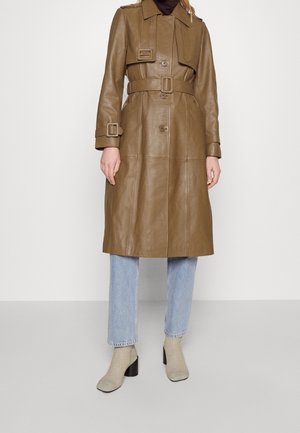 Woman wearing a knee-length brown leather trench coat with belt, light blue jeans, and beige heeled sock boots against a plain background.